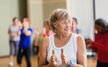 An active older woman in a white tank top and tawny shag haircut claps along in Enhance FItness class.