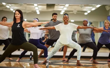 Women of different ages and ethnicities are in warrior pose in a YMCA studio with natural light.