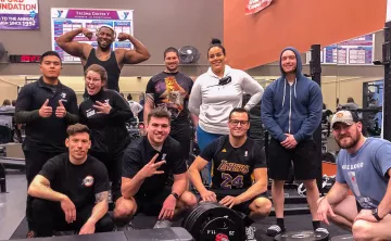 A group of adults huddle together and smile while inside the Tacoma Center YMCA weight room.