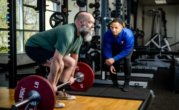 A man prepares to perform a deadlift with a Y staff member nearby in blue watching alongside for his form.