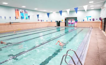 Indoor lap pool at the Tacoma Center YMCA with multiple swim lanes, lane dividers, and triangular flags overhead. Several swimmers use the lanes, with pool ladders and tiled deck visible along the edge.