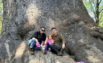 Sarah, her daughter Nora, and her husband sit together at the base of a large tree during a family outing.