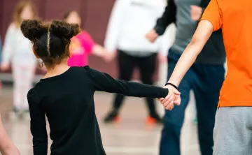A child holds hands with an adult while standing in a gymnasium, with several other people blurred in the background participating in a group activity.