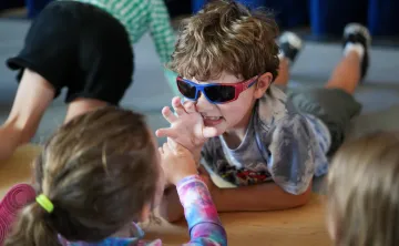 Children participating in an interactive educational activity indoors, with one child wearing colorful glasses while engaging with peers on a classroom floor during a youth learning program.