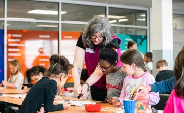 An instructor leans over a table to assist several children with an arts and crafts project at a YMCA classroom. Participants are seated around the table drawing and painting, with art supplies, cups, and paper spread across the workspace. Large windows and colorful wall displays are visible in the background.