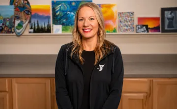 Michelle Wells, Arts Director, standing indoors in front of a display of colorful artwork and paintings. Wearing a dark YMCA-branded jacket over a black shirt, with light wood cabinetry visible below the art display.