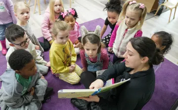 Early learning students gather on a classroom rug as an educator reads a picture book during group story time. Children sit in a circle around the reader in a bright early learning classroom with chairs and learning materials visible in the background.