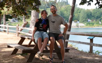 Alt text:  Aaron and Ryan sit together on a wooden picnic table at Camp Seymour beside the waterfront. Trees shade the area, with a wooden fence, calm water, and greenery visible in the background on a sunny summer day.