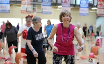 Adult participants take part in a group fitness class at a YMCA gymnasium. One participant in a bright pink sleeveless top exercises near the front of the group, with chairs arranged throughout the room for support. YMCA banners hang in the background above the activity area.