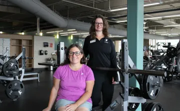 Julia poses with a YMCA staff member in the fitness center at the Gordon Family YMCA. Julia sits on a weight bench in front of a barbell while the staff member stands beside the bench. Strength training equipment and exercise machines are visible in the background.