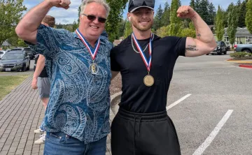 Two medal-winning participants flex their arms after the Haselwood Family YMCA Strongman Competition, standing outdoors in a parking lot following the community strength event in Silverdale, Washington.