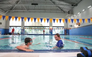 Young swimmer practicing skills during a private swim lesson with an instructor in an indoor YMCA pool. The pair are positioned near the pool edge, with lap lanes, blue and gold pennant flags, and large windows visible in the background.