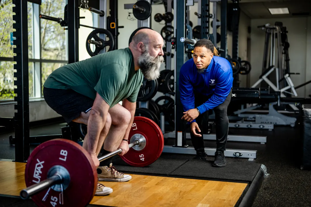 A man prepares to perform a deadlift with a Y staff member nearby in blue watching alongside for his form.